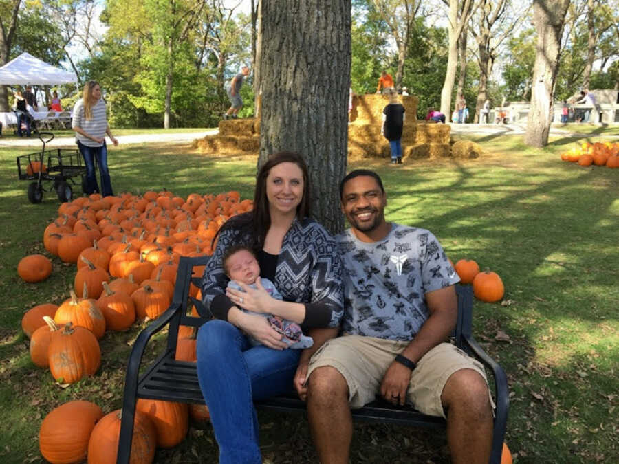 mom holds her son while sitting next to husband on bench at pumpkin patch