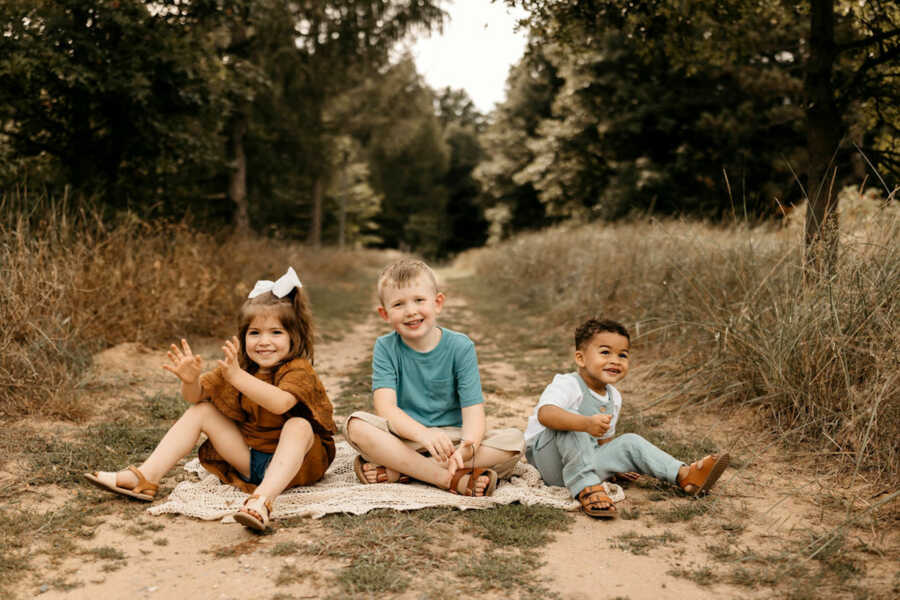three siblings sit on blanket on a path outside