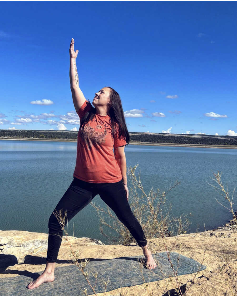 woman stands in front of lake doing yoga pose