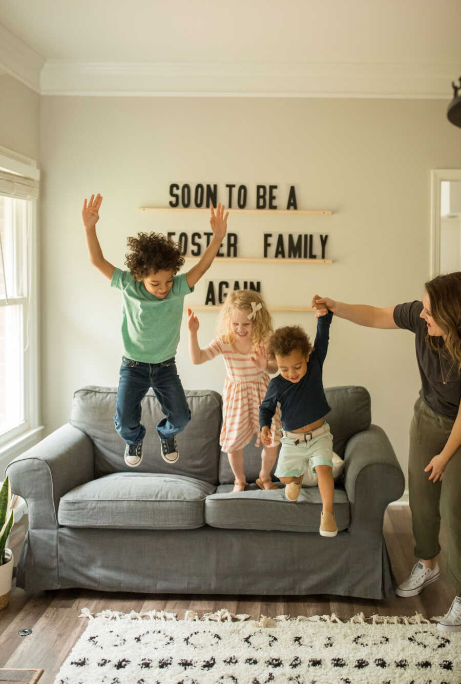 foster siblings jumping off grey couch with mother helping them play