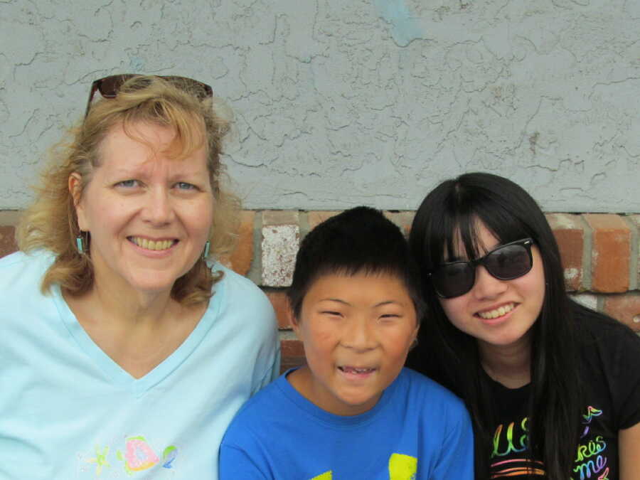 Una madre con su hija y su hijo con un trastorno del espectro alcohólico fetal sonriendo para tomar una foto afuera frente a una pared de ladrillos