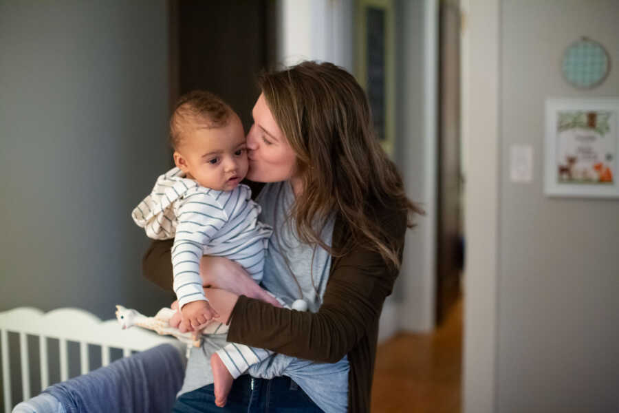 adoptive mother kissing adopted baby son that's wearing white and blue striped onesie 